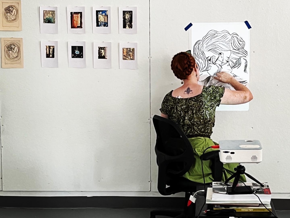 a person in a green blouse and skirt sits with back turned to the viewer and is drawing on a piece of paper taped to a studio wall, otherwise mostly bare save for a series of prints. 