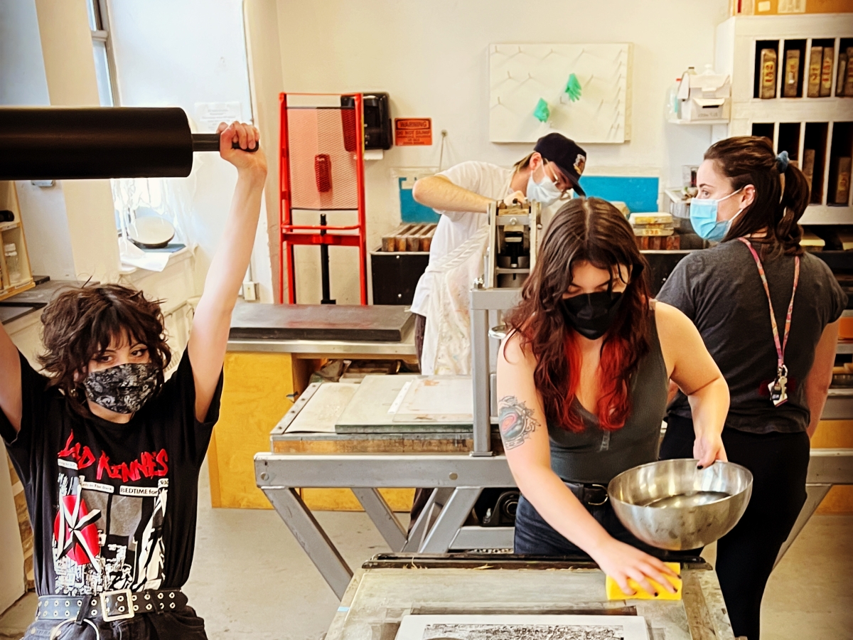 a group of students in the lithography studio. a person in the front left with a massive wallet chain and messy hair is holding a roller straight overhead. in the center, a person with long hair looks down and wets a stone with a sponge, a metal bowl in the other hand. a person in the far back is adjusting a press. all four people are wearing face masks. 