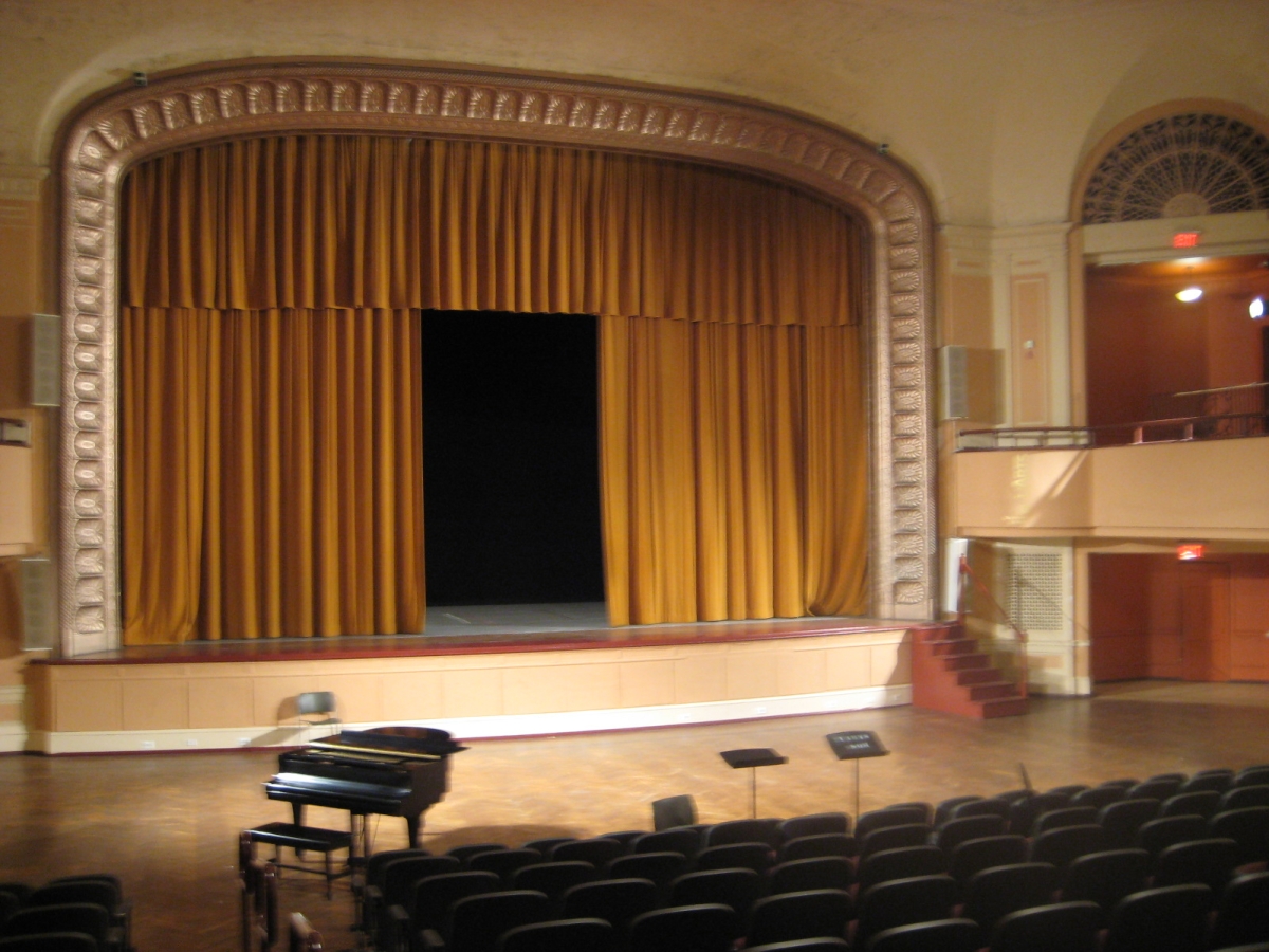 interior of levitt auditorium. the auditorium is empty, seen from the top of the central seating area. golden-yellow curtains are hanging across the large stage, with a large rectangular rift left open.  