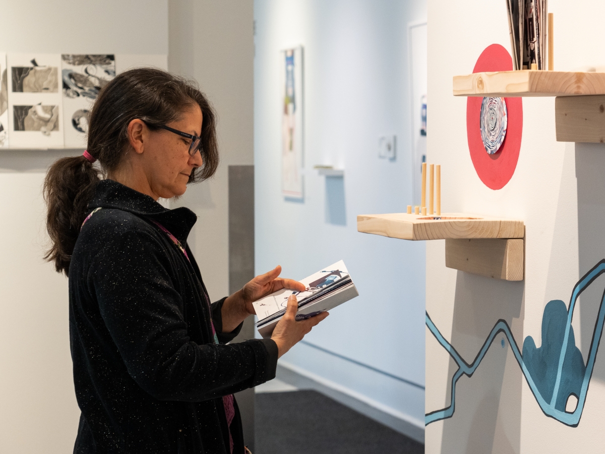 works on display at arronson gallery, with a person referencing a booklet at the left. the wall has small wooden shelves and blue zigzagging lines with a red illustrated animal skull