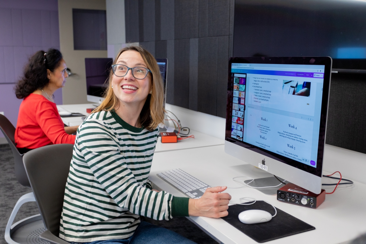 A smiling woman with glasses and a striped shirt is seated in front of an iMac looking over her shoulder. Another student with dark hair and a red shirt is visble at another computer beside her.