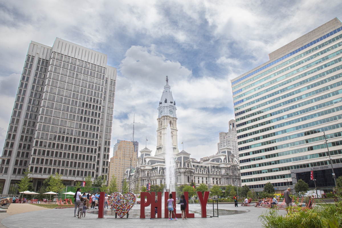 Some people are gathered around a public sculpture in a park that spells "I (Heart) Phily." A water fountain sits between the sculpture and two large buildings in the background with a view of the iconic City Hall building in Center City Philadelphia. It is a cloudy but bright blue sky.