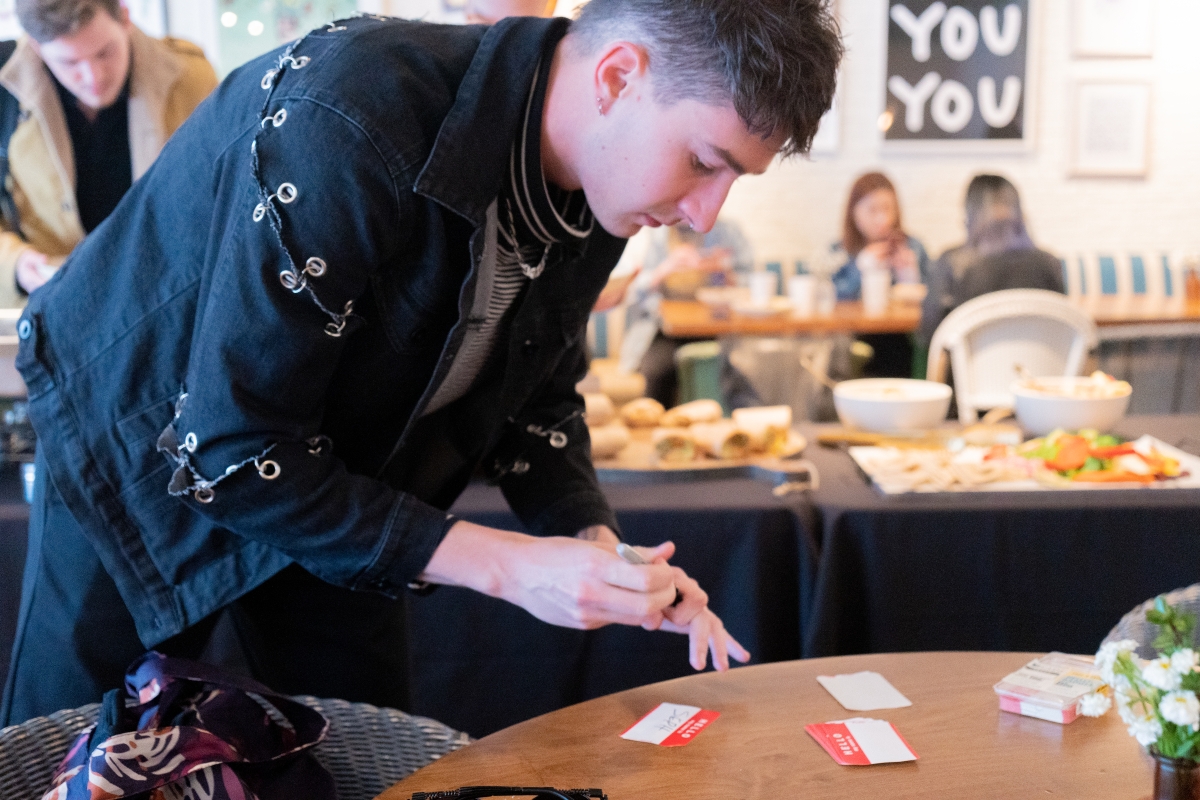 A person wearing a black denim jacket with rivets and clasps that hold various portions of the arms and shoulders of the jacket together has just written their name and is holding a marker, leaning over a table of nametags at a UArts Alumni function. In the background, a buffet and several tables are visible with figures seated across from each other.