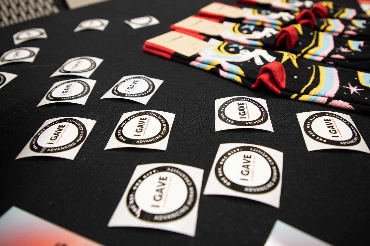 a table covered in black fabric has several circular stickers on backing paper scattered in the foreground. pairs of socks are lined up neatly in the background. The stickers have prominent text reading “I GAVE” and feature a ring around the outside. Most of the other text in the stickers is not legible, but UArts’ mission (“Advancing Human Creativity”) is aligned with the bottom-half of the ring and the Day of Giving motto (“One Day. One Gift.”) is aligned with the top. The socks are mostly black with colo