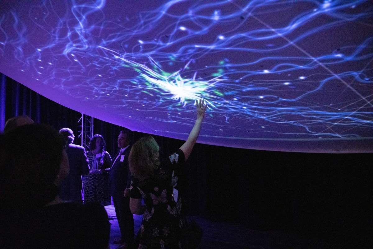 A large fabric blankets part of a dark room, curved in such a way that it resembles a planet or dome. A person in the foreground is reaching up to touch the fabric, which is illuminated and decorated with nebulous streaks and dots. The nebulous imagery is concentrated around the person's hand, presumably responsive to touch and motion. There are others visible standing and observing, lit dimly by the interactive display.