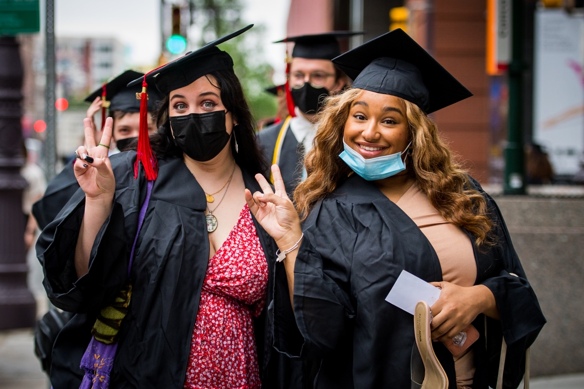 Two figures are side-by-side in the foreground of a group of four visible people who are all wearing robes and graduation caps with tassels. The people in the front are smiling for the camera, posing casually, with their hands forming peace signs. One is wearing a black surgical mask pulled up above their nose and the other has their mask pulled below their chin so the viewer can see their smile.