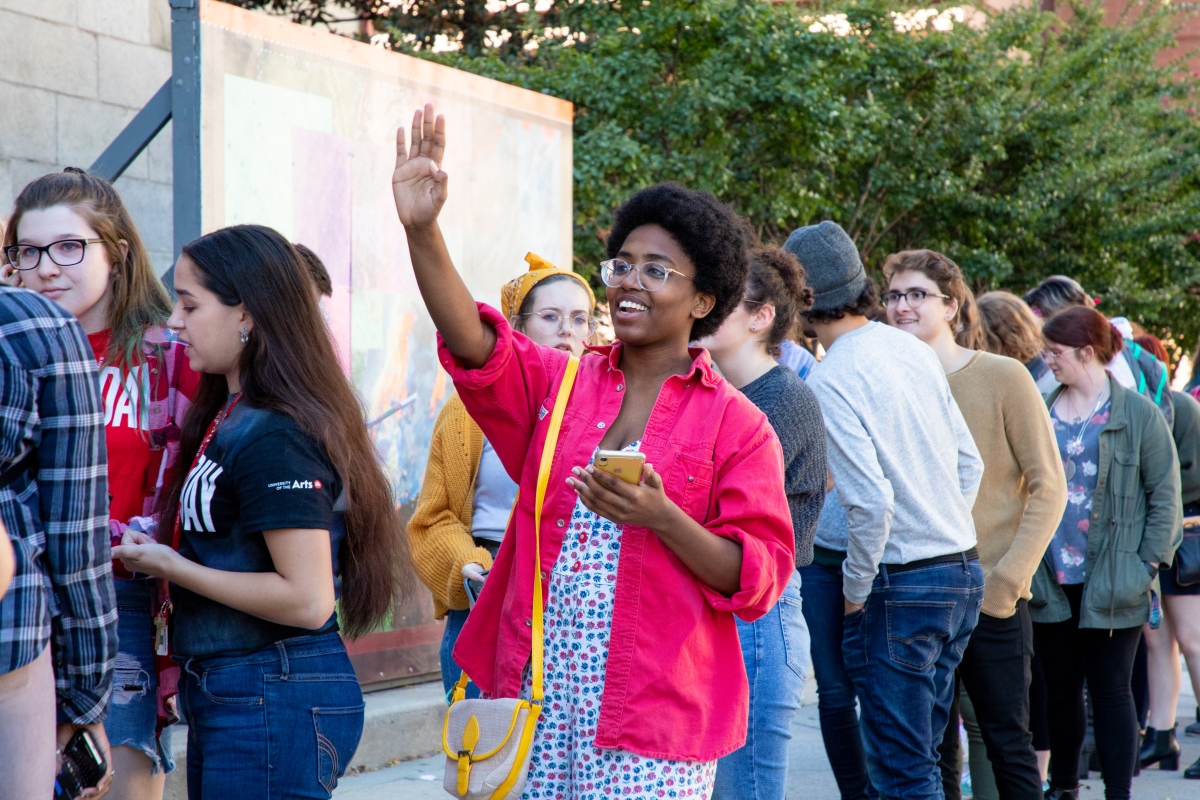 A person with dark skin and short, naturally styled dark hair and glasses wears a bright pink jacket over a colorful outfit underneath, standing among other people in a line outdoors. In one hand, they are holding what looks to be a phone. A bright handbag is slung over their other shoulder and they are raising their arm, smiling as though they are greeting a friend. The other people visible in the photo are queued by one or two people, talking to each other or looking off away from the camera. 