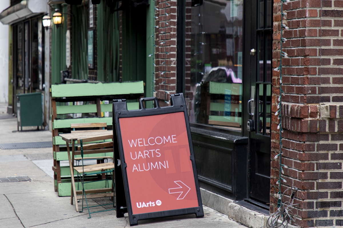 A clapboard sign that reads "Welcome UArts Alumni" with an arrow pointing toward a door. The sign is sitting on the sidewalk.