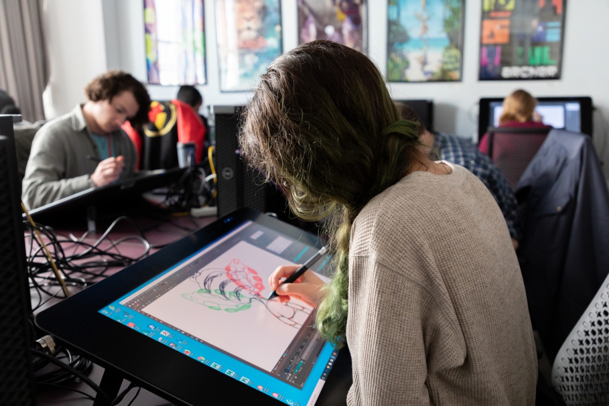 a person with long hair dyed green at the end is seen from their left shoulder in profile working on a digital drawing on a large wide tablet. in the background are additional people working on similar tablets or on computers with their backs turned toward the viewer. 