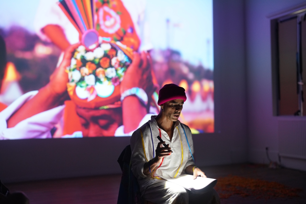 a person seated in a room illuminated by a projection against the back wall. the projection depicts from the nose up a person placing an ornate garment on their head in hues of old film. the person seated in the room is shining a flashlight ot a notepad in their lap. 