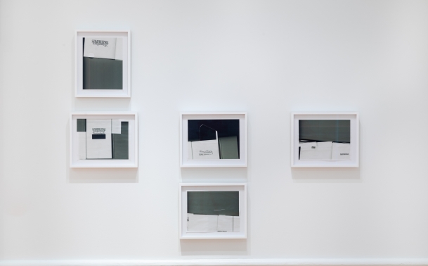 five grey scale collage studies in white frames hang on a white gallery wall. The collages are comprised of mostly abstracted compositions made from greyscale xeroxed pages from books, with some text fragments visible.