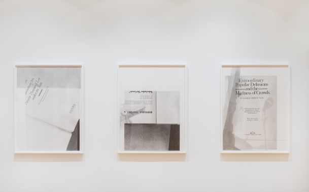 Three graphite drawings by Nyeema Morgan hang in white frames on a white wall. Each depicts the title page of a book with some fragmented text, white space, and darker grey shadowed areas included in the compositions.