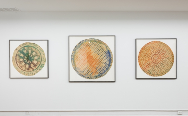 three framed works hang on a white wall. The composition of each work includes a white background, with a round multicolored central shape with various symbols and textures visible.