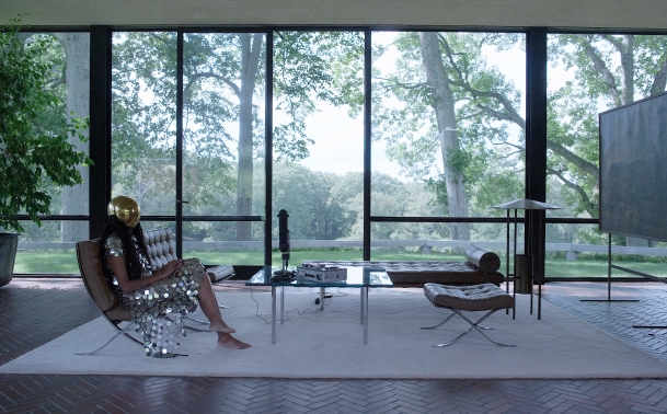 Digital film still showing a glass house and person wearing a gold helmet sitting on a couch