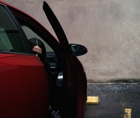 A photograph of a person inside a car, the car door is slightly open. The background is a concrete wall.