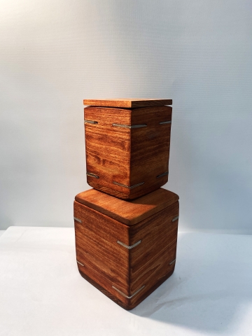 artwork by Feyisara Olalowo photographed against ablanket white background. two boxes made of red-stained wood with horizontal grain are shown with a smaller one resting on top of the other. the corner edges each have two gray slits 