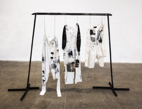 Meg Meehan's artwork consisting of a black scaffolded stand with three white garments hanging from the cross bar, each embroidered and printed with cryptic textural black designs