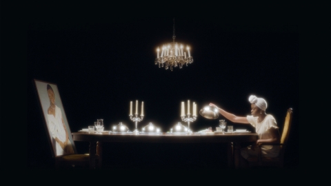Still from xenia matthews film Ourika!. in a completely black scene, a single long dining table is liminated brightly and seen in square profile, lengthwise. at the right end is a person in a white dress with white headscarf leaning forward to lift the silver lid off of a serving platter, revealing no food. there are several more covered silver platters. at the left end is a portrait of a person in aw hite dress. a chandelier is suspended in blackness above the table. 