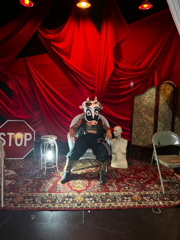 santi seated in a white wicker chair wearing snakeskin cowboy boots with a large goofy cow head mask. the back wall is all red drapes, with a white bust to the right of the chair and a stop sign to the left 
