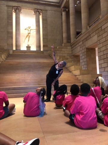 santi standing in a grand marble chamber against a backdrop of stairs with a golden statue holding a bow at the top. children in pink tshirts are seated on the floor looking at Santi, who is leaning and pointing and looking directly up at the ceiling. santi is wearing all black. 