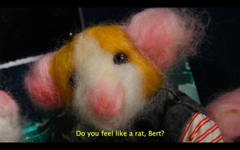 a still from animation work by katarina mortko. a little rodent puppet made of felt has pink ears and black beady eyes is wearing a tiny denim vest and a striped shirt. the critter has yellow subtitles below it, speaking Do you feel like a rat, Bert?