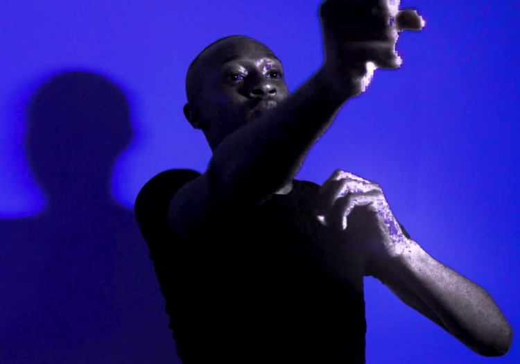 still from Xenia Matthews film Stroke. a person rendered in black and white in a black tshirt motions with both arms while standing against a deep blue background. the person is rendered in a reduced fidelity, as if little snippets of data are missing.  