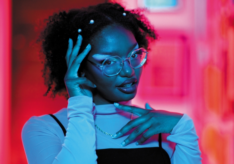 Headshot of Xenia Matthews. Matthews is illuminated in blue lighting and is standing in an out of focus hallway bathed in pink light. She is wearing a white turtleneck with a black thin-strapped tank top over it, with clear acrylic glasses on her face. Matthews is looking at the viewer with her head angled slightly to the side and down. one hand is raise to her chest, one to her temple. there are pearls in her hair.  