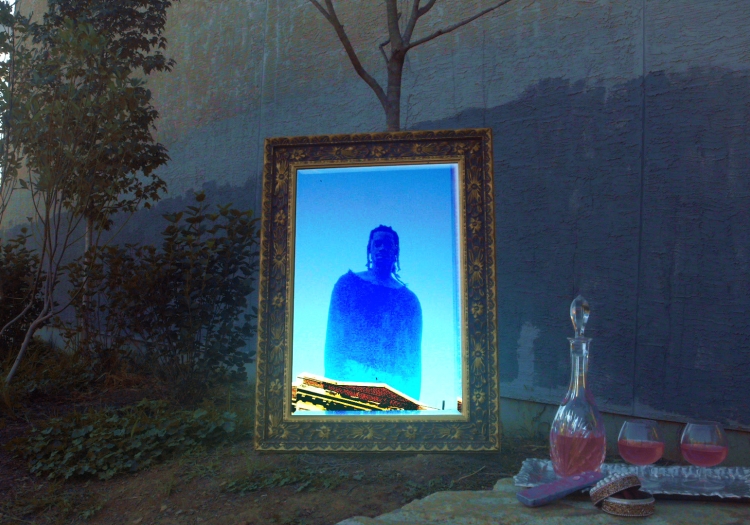 Still from Xenia Matthews' film A Few Things I'm Beginning to Understand. An ornate mirror stands in an almost surreal landscape by a mural-painted wall, leaned up against a bare small tree. In the mirror is the reflection of a person, rendered in a blown-out blue hue, as if the camera was over-saturated. 