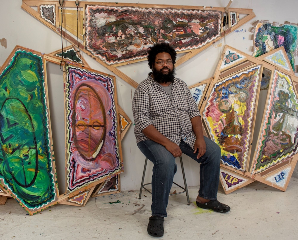 Artist Mike Cloud sits on a still in front of his artwork in his studio wearing jeans and a plaid shirt