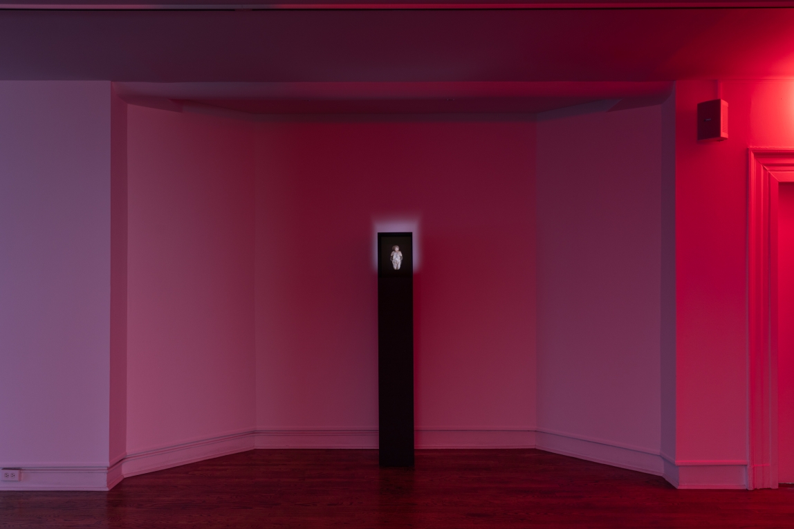 An installation shot of Abbey Williams' work shows a gallery space illuminated with red light with a black pillar at the center. The black pillar shows a digital image of a statue 