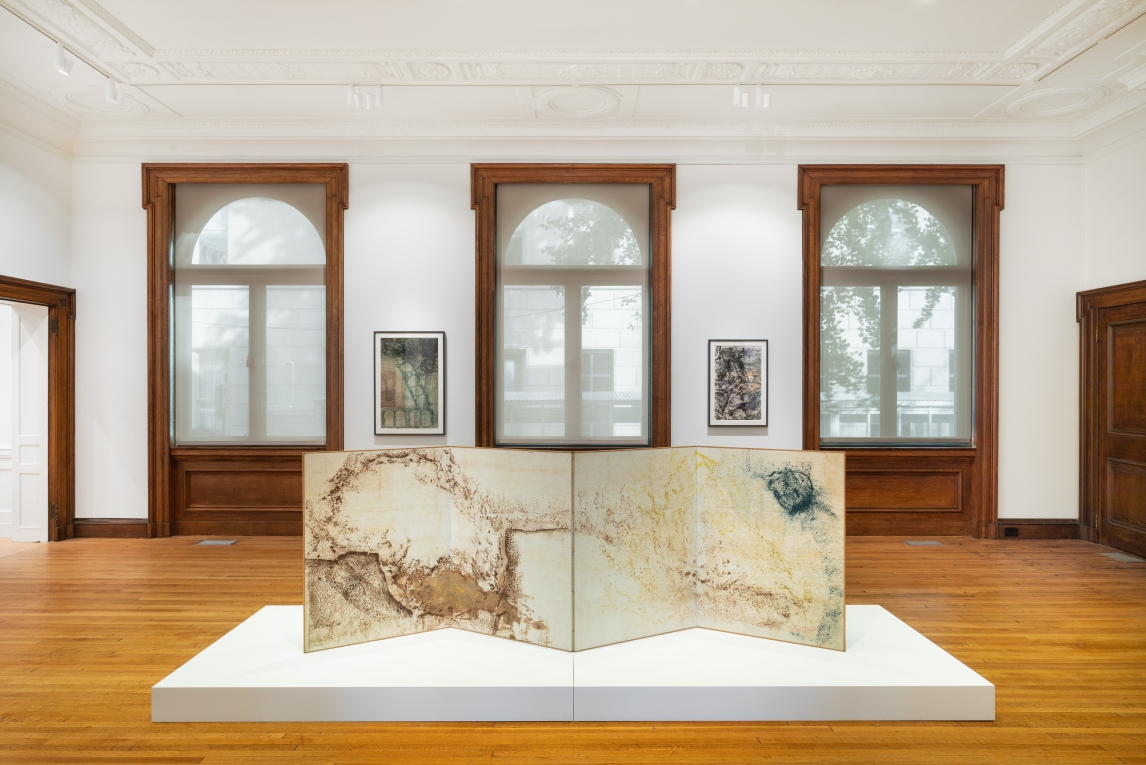 a gallery installed with Sari Dienes work. A large multipanel abstract work on a white pedestal is in the center of the image. In the background are three floor ceiling windows with two framed works hung between them