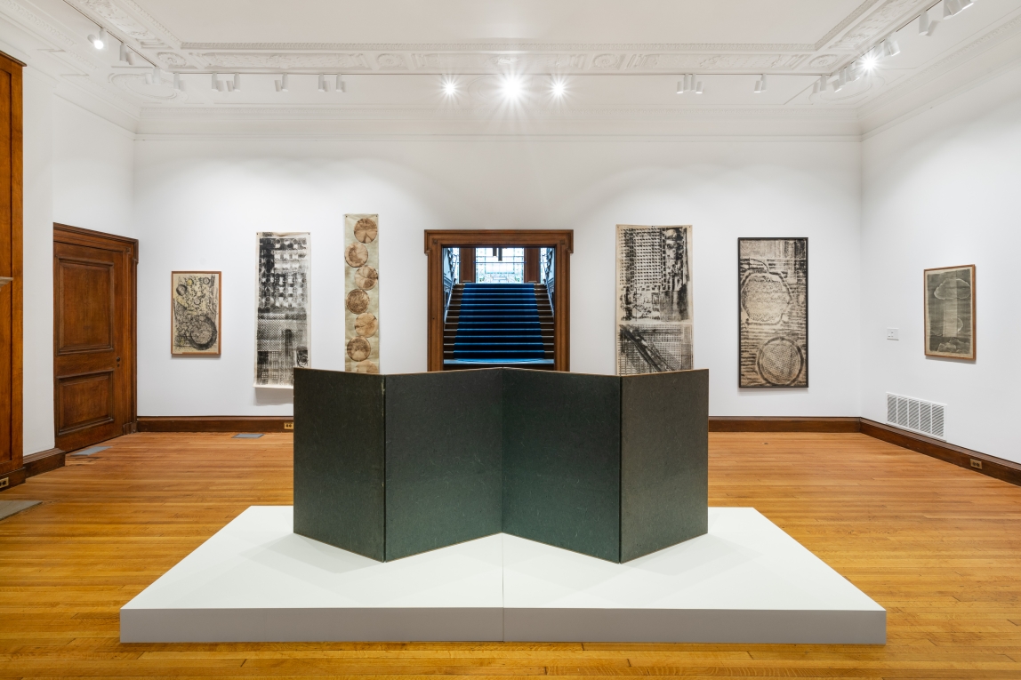 an gallery installation shot featuring a large multipaneled work on a white pedestal in the center of the image. In the background work of a variety of sizes and media hangs along a large white wall