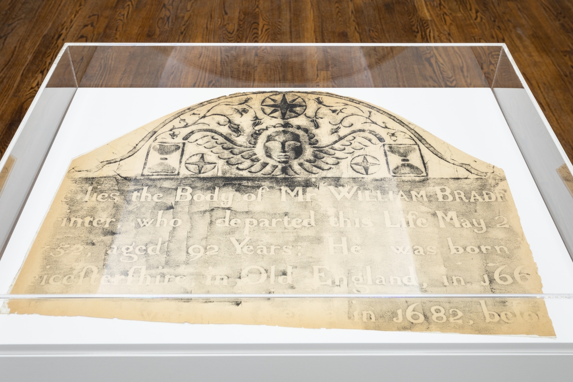 a large ink rubbing of a gravestone sits on a white pedestal encased in plexiglass.