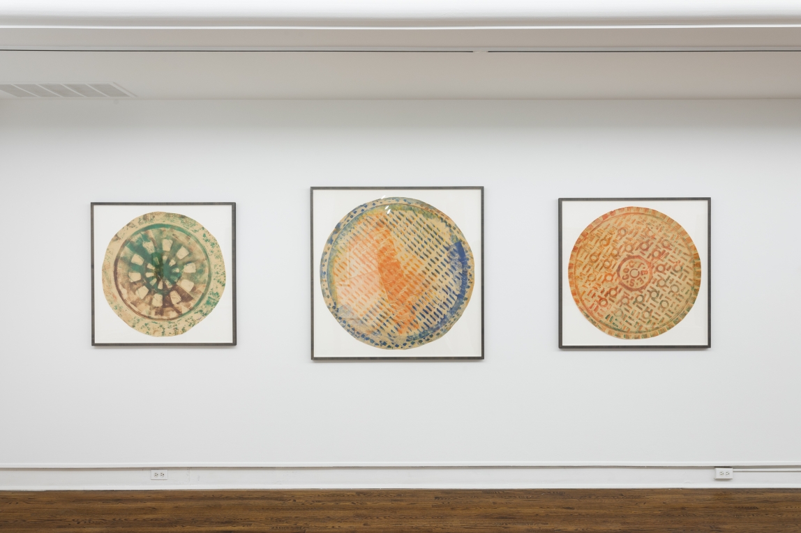 three framed works installed in a horizontal row on a white wall. Each composition includes a round mutlicolored central shape, with various textures and symbols, against a white background.