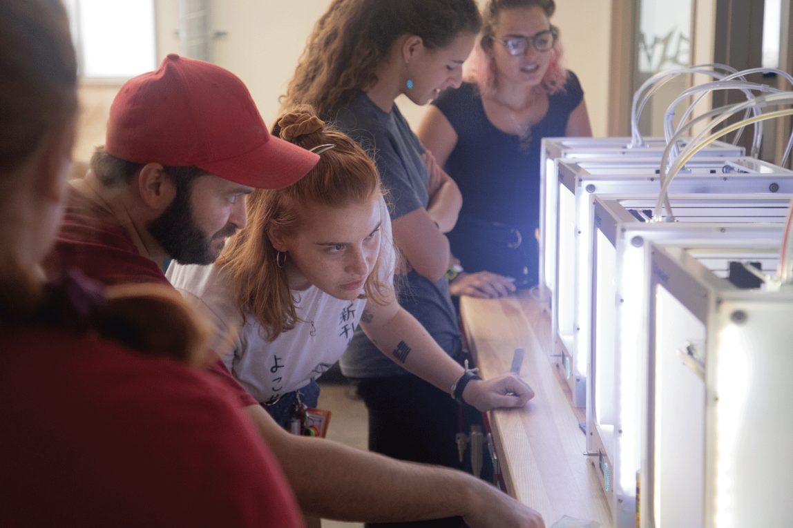 A tight shot looking over the shoulder of one student at a bearded man in a red hat and a student who is leaning down looking intently into a row of small, tabletop 3-d printers. Two other students are standing in the background.