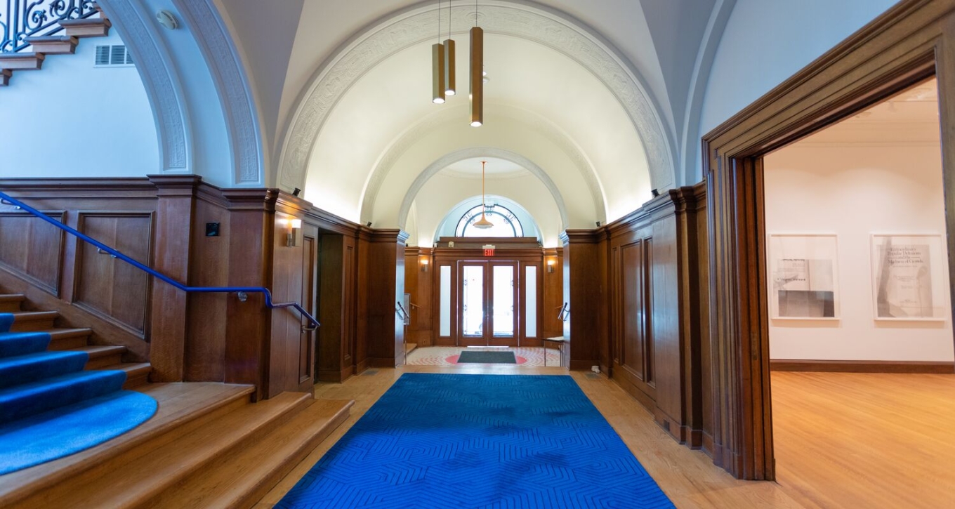 In an ecclectic mix of modern and traditional styles, an entryway is depicted with a curvilinear arches at the end of a hallway. Bright blue carpet runs down the hall and up the stairs to the left. The stairs also feature a bright blue banister. Warm, natural wooden paneling wraps around the space, and the archways and ceilings are bright white. A cylindrical chandelier hangs in the center of the space. Just off of the hallway to the right, an entrance to a gallery space.