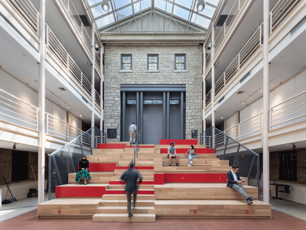 Several figures sit and walk on an asymmetrical staircase in natural wood decorated with red. The steps are central in a light-filled interior courtyard with high, glass ceilings and three "catwalk" like floors on either side of a neoclassical stone wall that has three windows and a large portal painted in gray with columns running across.
