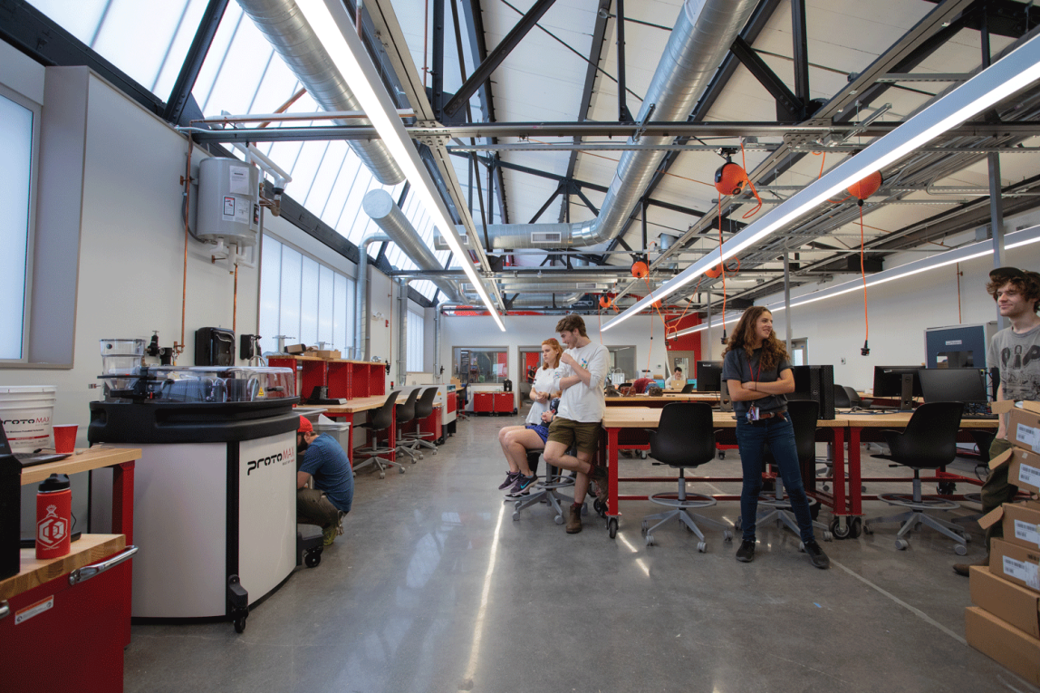 Students are gathered in a light-filled, modern and rather industrial looking space, filled with work tables, chairs and equipment. Exposed ducts, dog-house style windows and bright lights adorn the vaulted ceiling. Power strips, red accents and a glossy concrete floor complete the space.