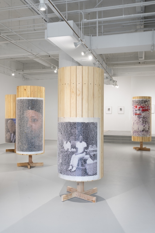 Four wooden columns in gallery, with posters of people on them, covered in tacks