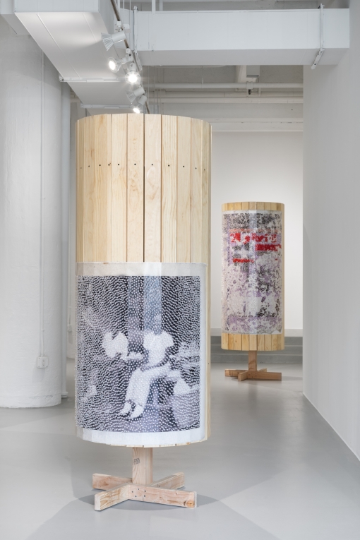 two wooden columns with images covered in tacks, one in foreground with seated figures, one in background with red and clear tacks