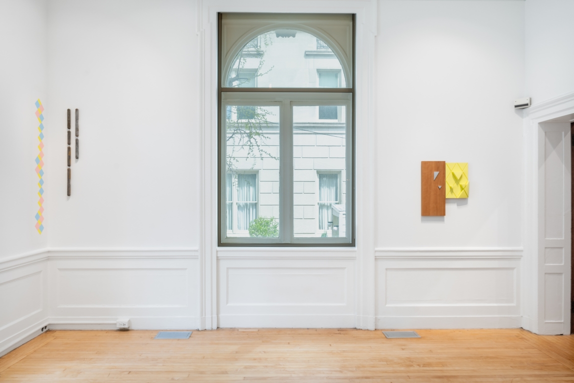 Installation view of two wall sculptures on either side of a large window, one made of metal, the other plaster and wood