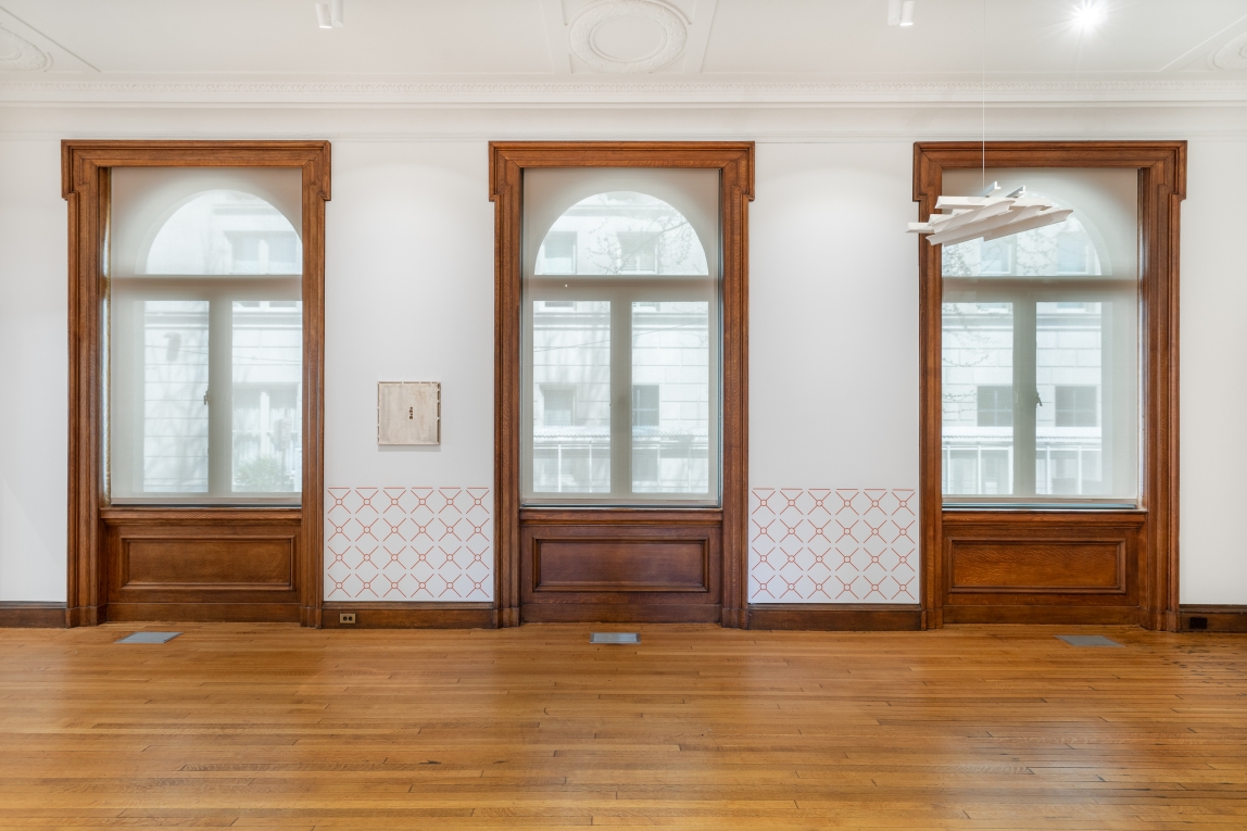 Installation view of Gallery B with wall decals in-between three large windows and a hanging white grid-like sculpture in foreground