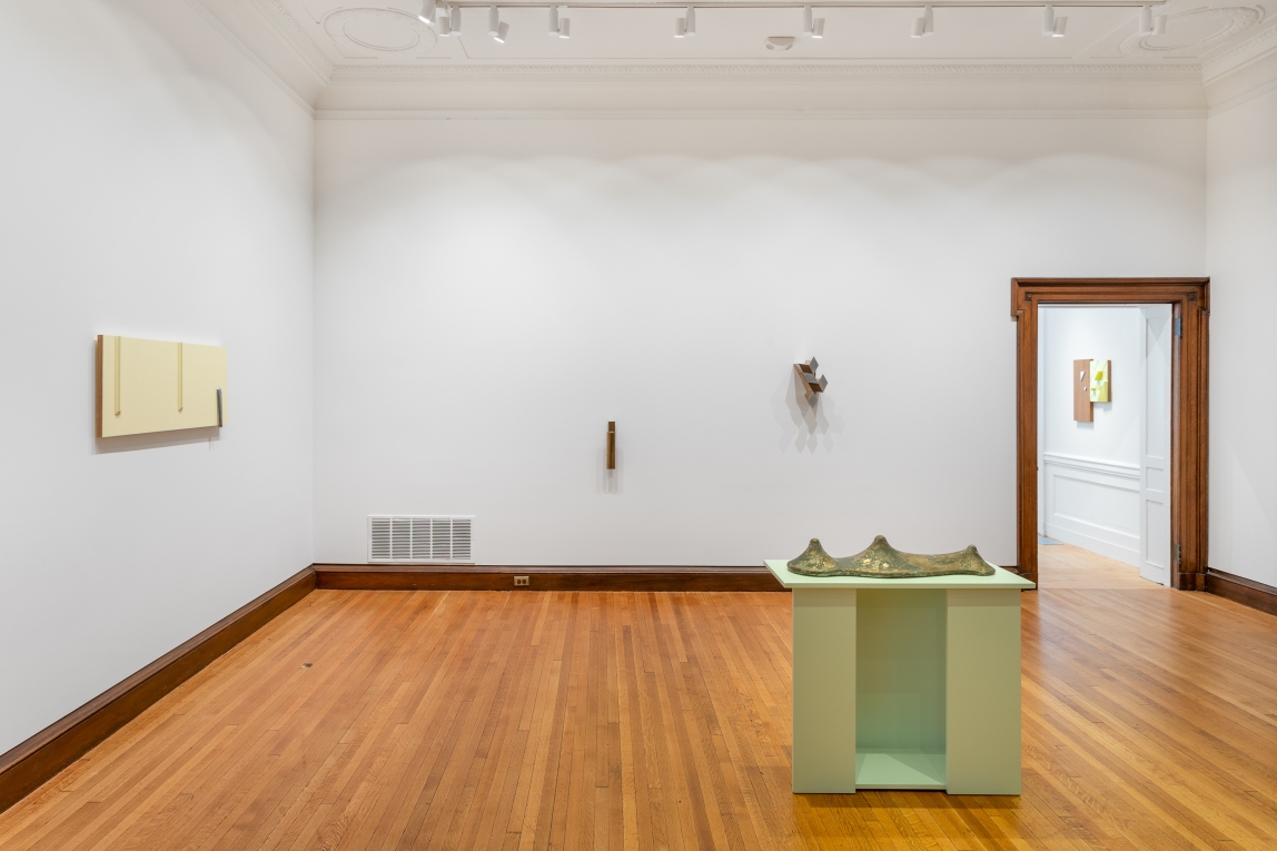 Installation view of 4 sculptures: a yellow plaster/cast work on one wall, two separate metal works on the back wall and a green pedestal with a bronze work on floor in foreground