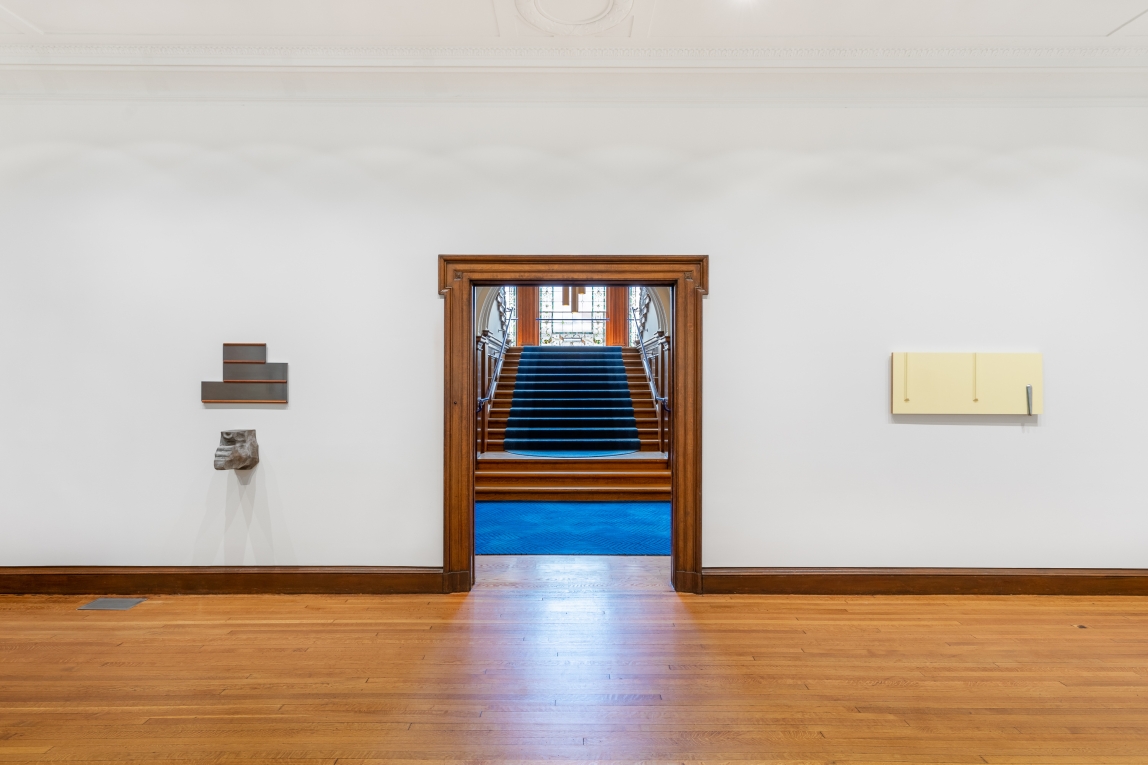 installation image with one metal wall sculpture to left and yellow cast and metal wall work to right bisected by doorway leading to grand staircase