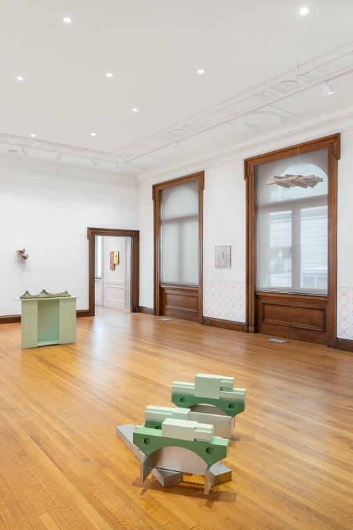installation view of room with sculptures: a green cast and metal work on floor, metal work on pedestal in mid ground, white cast work hanging from ceiling and two metal works on walls
