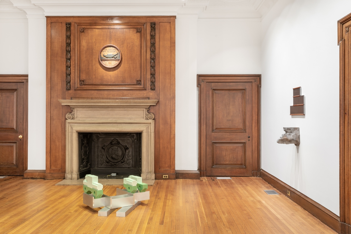 Installation image of a floor sculpture oof casting and metal and a wall sculpture of bronze and other metal with fireplace in background