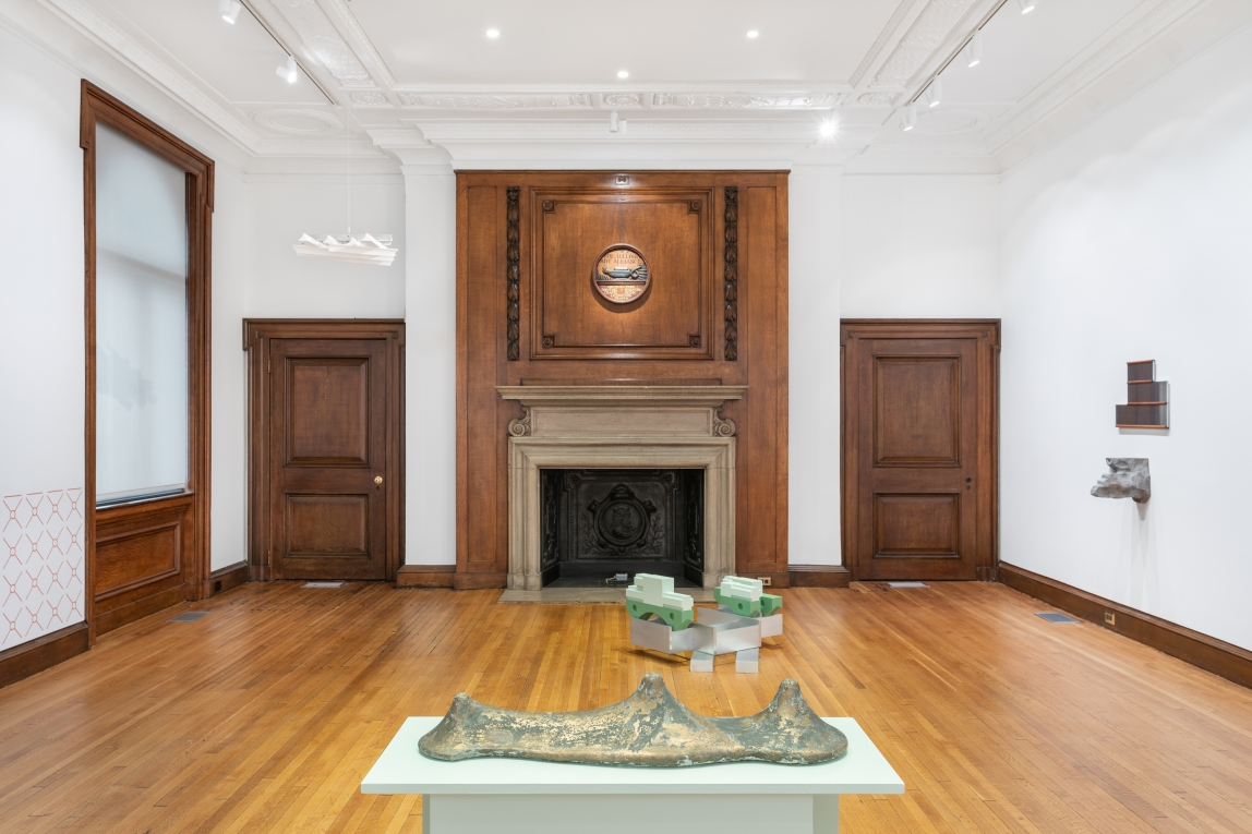 Installation image of several sculptures in a large white room with dark wood trim and fireplace 