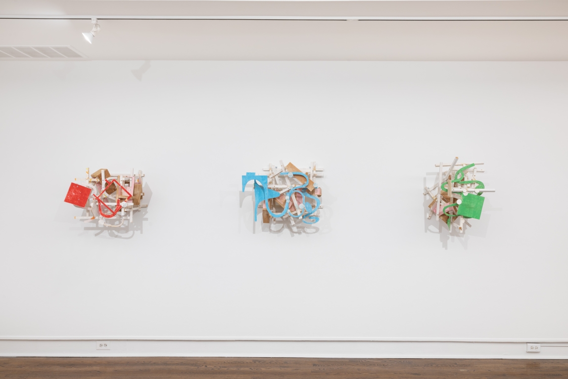 Installation view of three brightly colored assemblages hanging in a row on the wall