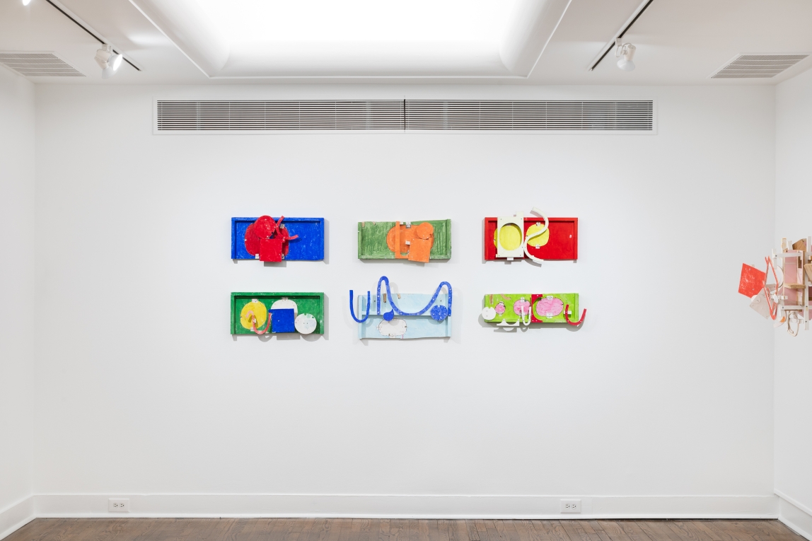 Installation view of six brightly colored flat assemblage works hanging in two rows on the wall with one other assemblage work on the right