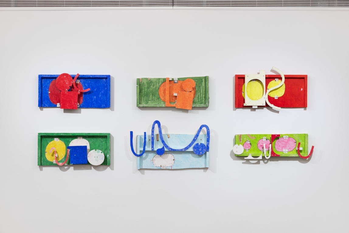 Installation view of six brightly colored flat assemblage wall sculptures,  in two rows
